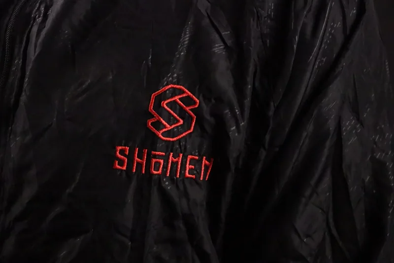 Pormenor do logótipo e da palavra Shomen a vermelho no corta-vento preto